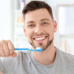 Man in grey shirt smiling while brushing his teeth with blue toothbrush