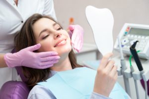 Woman with brown hair in dental chair holding up a mirror with a dentist wearing purple gloves behind her